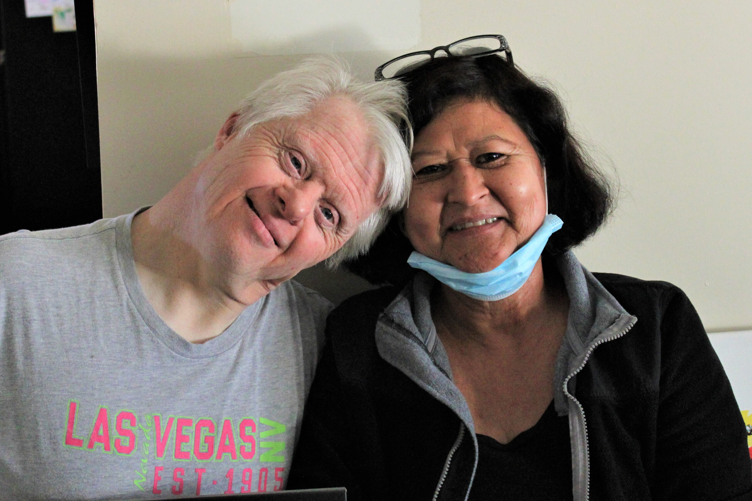 A resident at one of the homes The Campbell Center manages is smiling and tilting his head of white hair towards his caregiver, who is a woman smiling, with dark brown hair and a mask on her chin.