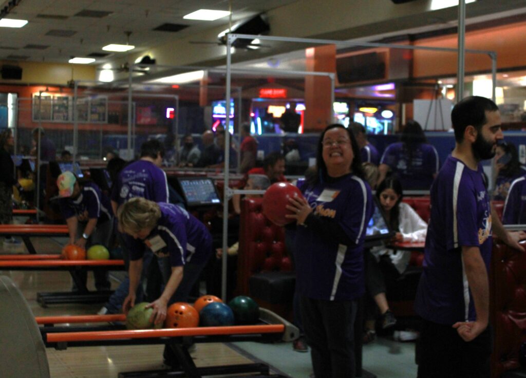 Bowlers in purple Bowlathon event shirts getting in position to throw their bowling ball. One in the center is a woman with short brown hair who looks at the camera holding her red bowl close to her smiling at the camera.