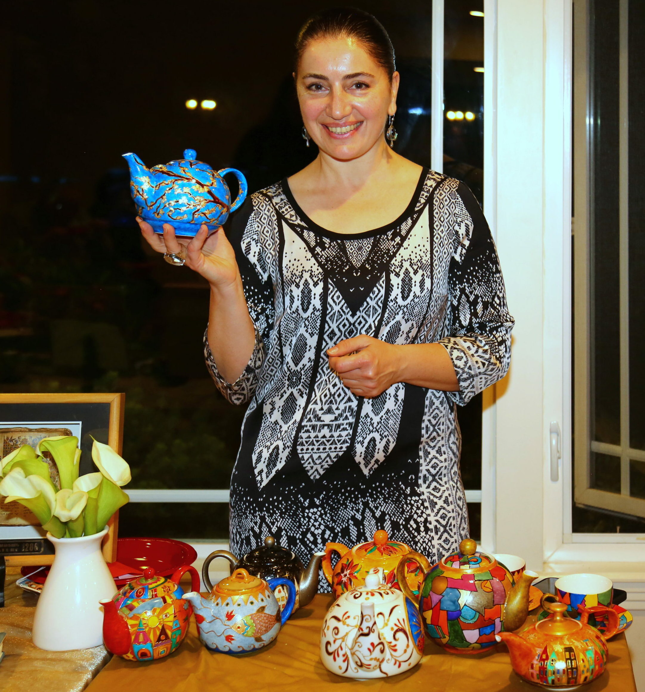 Tamara smiling holding up a blue teapot she made above selling colorful pottery smiling with a black and white dress on.