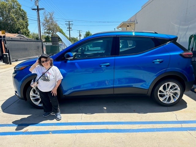 Short woman standing next to blue car smiling.